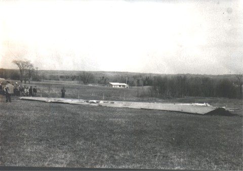A large section of wing wreckage in a farm field, with onlookers gathered.